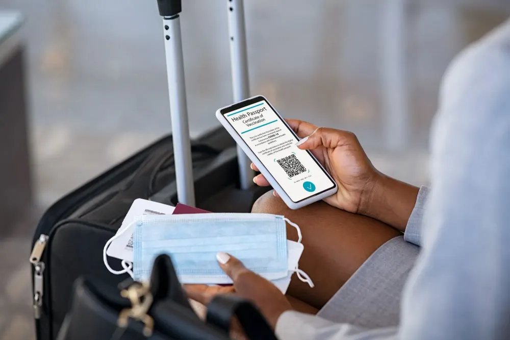 Woman sitting by her luggage, holding a facemask and a phone showing a COVID-19 Health Passport